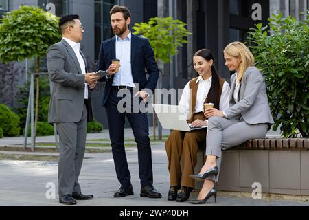 Drei Geschäftskollegen in intelligenter Kleidung diskutieren informell vor einem Bürogebäude mit Kaffeetassen und einem Laptop. Stockfoto