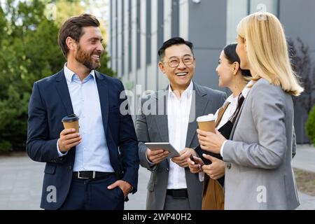 Vier fröhliche Kollegen in Geschäftskleidung tragen Kaffee zum Mitnehmen und führen ein freundliches Gespräch vor einem modernen Büro. Stockfoto