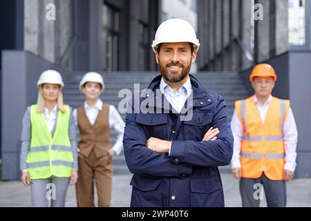 Professioneller, männlicher Architekt lächelt in Schutzhelm mit diversen Konstruktionsteams in Warnwesten. Stockfoto