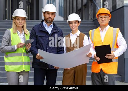 Vier Bauprofis in Schutzhelmen prüfen den Plan außerhalb des modernen Gebäudes, konzentriert und entschlossen. Stockfoto