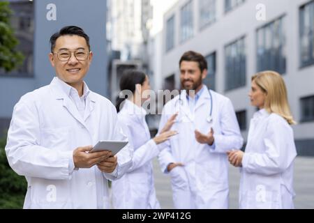 Selbstbewusster asiatischer männlicher Arzt, der ein Tablet hält, lächelt in die Kamera mit dem medizinischen Team, das sich hinter ihm in der Diskussion engagiert. Stockfoto