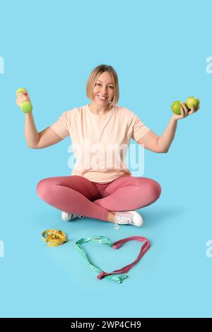 Hübsche junge Frau mit Maßband, Äpfeln und Hantel auf blauem Hintergrund. Gewichtsverlust-Konzept Stockfoto