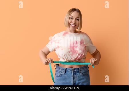 Hübsche junge Frau in lockerer Jeans, die ihre Taille auf beigefarbenem Hintergrund misst. Gewichtsverlust-Konzept Stockfoto