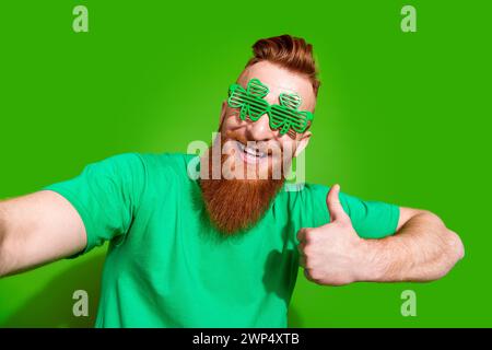 Foto eines attraktiven optimistischen Mannes mit Ingwerbart in Kleebläsern mit Daumen nach oben Make Selfie isoliert auf grünem Hintergrund Stockfoto