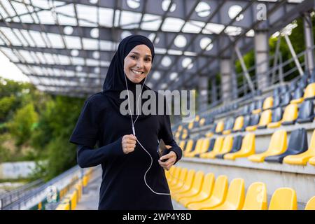 Positive islamische Frau in Hijab und sportlicher Kleidung, die Übungen mit kabelgebundenen Ohrhörern macht, die mit dem Mobiltelefon verbunden sind. Lächelnde Dame, die gerne Zeit im Stadion verbringt und das Laufen zur Musik übt. Stockfoto