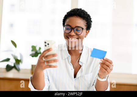 Online-Shopping. Positive junge afroamerikanische Frau in smarter Casual Shire, die Smartphone und Kreditkarte hält, im Internet Einkäufe tätigt, Geld online überträgt Stockfoto