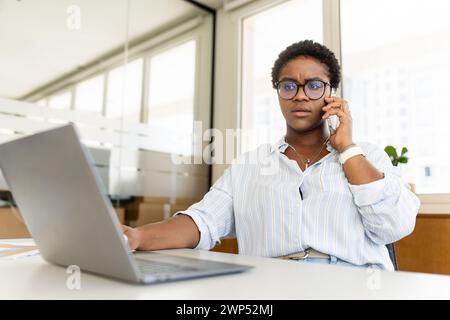 Besorgte afroamerikanische Büroangestellte, die auf dem Smartphone im Büro sitzt, eine Frau, die unangenehme Telefongespräche mit dem Vorgesetzten führt Stockfoto