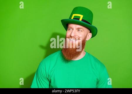 Foto von funky positivem Mann mit Ingwerbbart gekleidetem grünem T-Shirt Kopfbedeckung Blick auf Angebot leeren Raum isoliert auf grünem Hintergrund Stockfoto