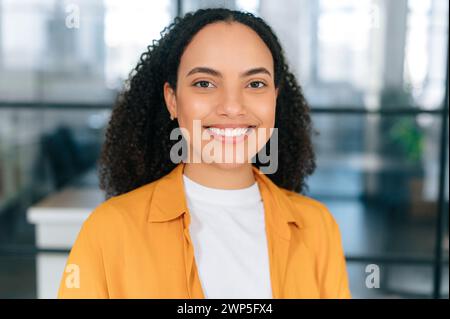 Nahaufnahme einer wunderbaren positiven, selbstbewussten brasilianischen oder hispanischen jungen Frau mit lockigen Haaren in einem orangefarbenen Hemd, steht im Büro, schaut in die Kamera und lächelt glücklich. Weibliches Porträt Stockfoto