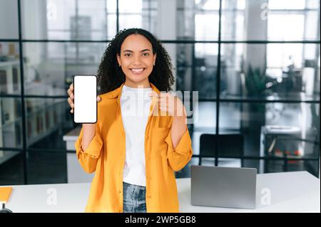 Niedliche positive, multirassische junge Frau, die im Büro steht, Smartphone mit leerem weißen Mock-up-Bildschirm für Werbung oder Ansage hält und mit dem Finger darauf zeigt, schaut und lächelt in die Kamera Stockfoto