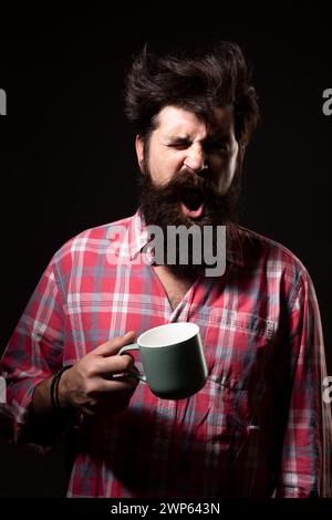 Mans hält eine Tasse Kaffee und Gähnen auf Schwarz. Morgentee. Gähngesicht. Mann mit Teetasse. Hipster-Mann gähnt mit einer Tasse Kaffee. Bärtiger Mann Stockfoto