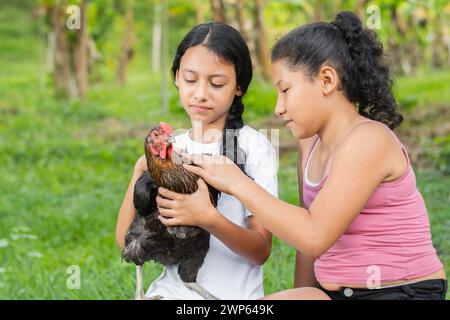 Zwei Latina-Bauernmädchen, die ein schwarzes Huhn halten, es streicheln und es ansehen Stockfoto