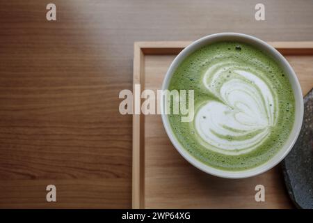 Ein schaumiger und cremiger Matcha-Latte mit Latte Art, serviert in einer Keramikschale zusammen mit einem Stück mehrschichtigen Matcha-Kuchen, präsentiert auf einer Keramikplatte. Stockfoto