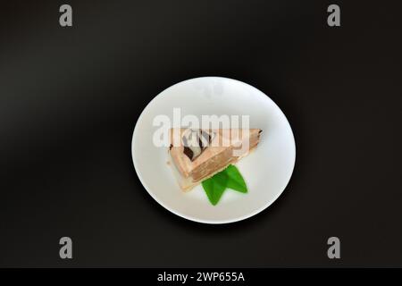 Ein Stück cremiger Käsekuchen mit Minzblättern auf einer weißen runden Keramikplatte auf schwarzem Hintergrund. Draufsicht, flach. Stockfoto