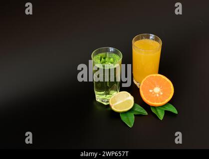 Zwei große Gläser mit Zitrussäften in einer Reihe auf schwarzem Hintergrund, neben reifer Orange und Limette mit Blättern. Nahaufnahme. Stockfoto