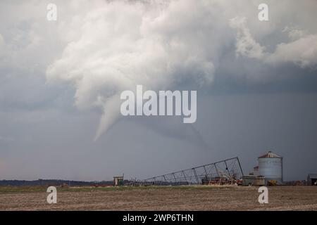 Ein weißer Tornado hängt unter einer Sturmwolke über ländlichem Ackerland mit landwirtschaftlichen Gebäuden und Ausrüstung im Vordergrund. Stockfoto