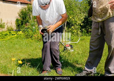 Ein Techniker mit verbandelten Handverletzungen repariert den Trimmer, stellt die Kopfspule des Trimmers ein und hilft seinem Freund im Schutzanzug beim Rasenmähen Stockfoto