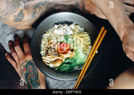 Eine Schüssel Nudeln mit Fleisch und Gemüse und Essstäbchen an der Seite. Die Schüssel wird auf einen Tisch gestellt, und der Arm einer Person ist im Hintergrund sichtbar Stockfoto