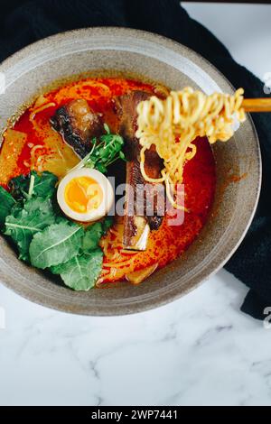 Eine Schüssel Ramen mit einem Stück Ei darin. Die Nudeln sind lang und die Brühe rot Stockfoto