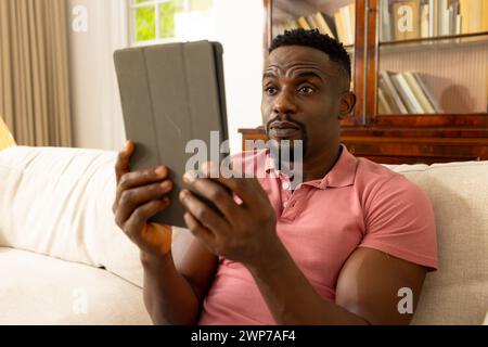 Ein Afroamerikaner in einem pinken Poloshirt sieht von einem Tablet zu Hause überrascht aus Stockfoto