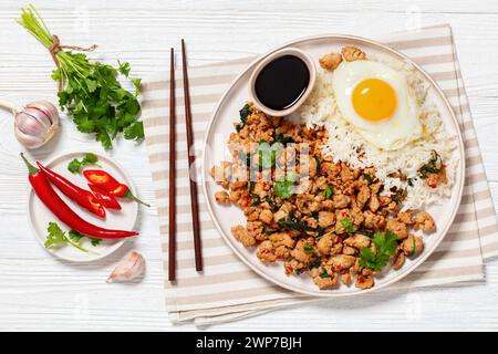 Pad Krapow Gai, Pad Gaprao, Pad kra Pao, Basilikum-Hühnchen-Rührbraten mit Reis und Spiegelei auf Teller mit Essstäbchen auf weißem Holztisch, thai-Küche, Stockfoto