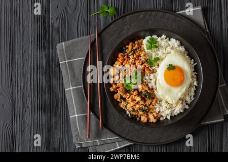 Pad Krapow Gai, Pad Gaprao, Pad kra Pao, Basilikum-Hühnchen-Rührbraten mit Reis und Spiegelei auf Teller mit Essstäbchen auf schwarzem Holztisch, thailändische Küche, Stockfoto