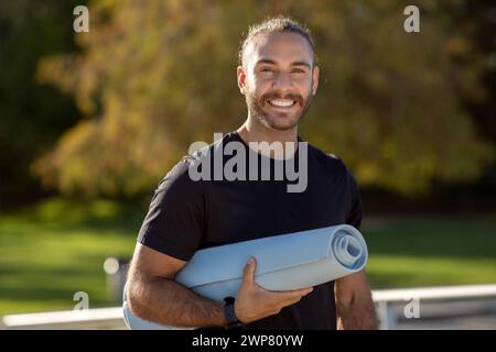 Glücklicher Sportler posiert selbstbewusst in Aktivkleidung und hält die Matte draußen Stockfoto