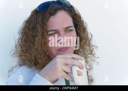 Attraktive, lockige, blauäugige Frau, die nachdenklich mit einer Tasse Kaffee in der Hand ist Stockfoto