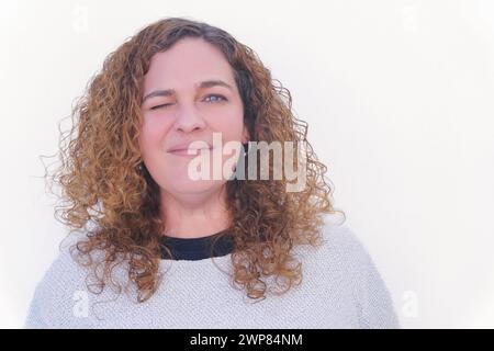 Schöne blonde Frau mit lockigen Haaren und blauen Augen in weiß, lächelnd in die Kamera und zwinkernd Stockfoto