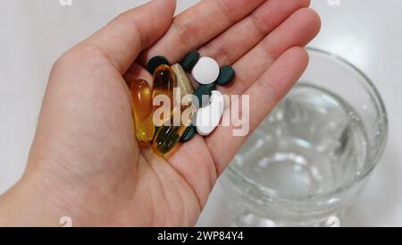 Hand hält verschiedene Ergänzungspillen und Kapseln. Mit einem Glas Wasser im Hintergrund. Stockfoto