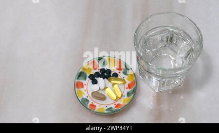 Ein kleiner Teller mit verschiedenen Nahrungsergänzungsmitteln Pillen und Kapseln. Auf einer Marmoroberfläche, mit einem Glas Wasser neben einem Teller. Stockfoto