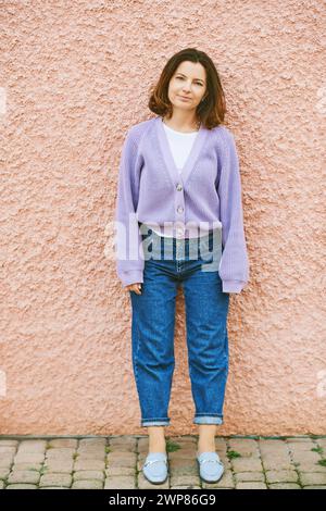 Außenporträt einer hübschen 40 - 45-jährigen Frau, die sich auf eine pinkfarbene Wand stützt, lila Strickjacke, Frühlingsstimmung und Farben trägt Stockfoto