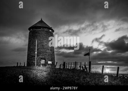 Ein Schwarzweißfoto eines alten Windmühlenturms am Meer bei Nacht Stockfoto