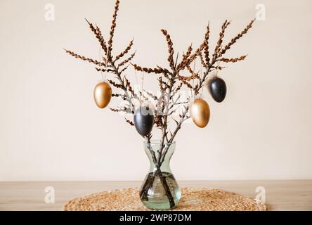 Baumzweige in Vase mit farbigen Ostereiern, die an der Schnur hängen. Schöne warme Beige-Erdtöne. Minimaler Osterschmuck. Stockfoto