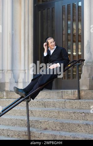 In der Pause sitzt ein junger orthodoxer jüdischer Junge auf einem Geländer und interagiert mit seinen Klassenkameraden. In Brooklyn, New York, März 2024. Stockfoto