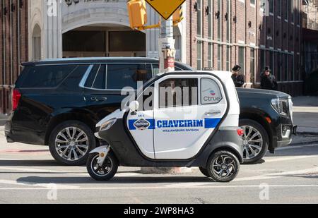 GROSS, KLEIN. Ein großer Cadillac Denali parkte hinter einem kleinen dreirädrigen Chaverim-Minifahrzeug in Williamsburg, Brooklyn, New York. Stockfoto