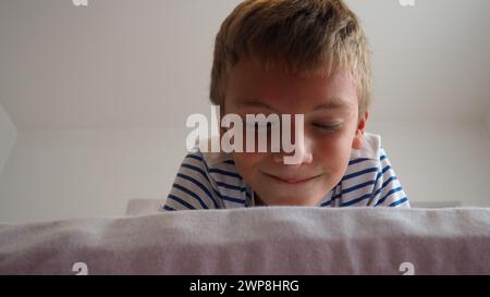 Der süße kleine kaukasische Junge, 10 Jahre alt, liegt zu Hause auf dem Bett im Schlafzimmer und lächelt lächelnd, schielend und schließt die Augen. Ich erwarte eine Überraschung Stockfoto