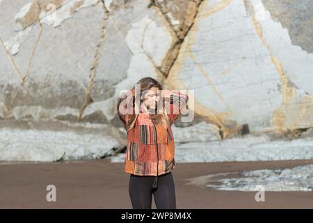 Eine Frau, die eine Jacke mit Patchwork-Design trägt, steht am Strand. Die Jacke ist rot und schwarz, und die Frau blickt in den Himmel. Konzept Stockfoto