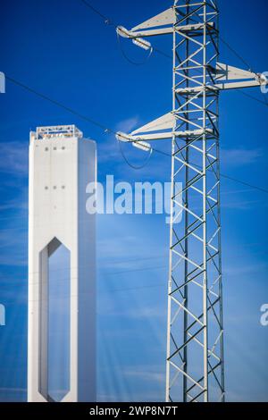 Hochspannungsleitungen, die neben einer hochaufragenden Solaranlage unter blauem Himmel verlaufen. Stockfoto