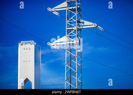 Hochspannungsleitungen, die neben einer hochaufragenden Solaranlage unter blauem Himmel verlaufen. Stockfoto