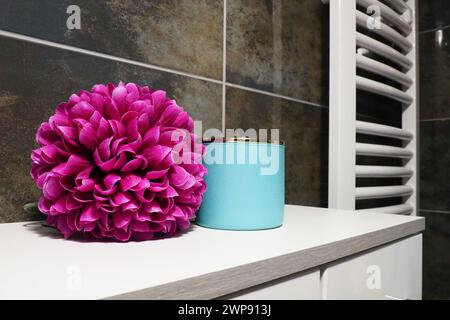 Dekorelemente im Badezimmer. Ein blaues Glas Creme und eine rosafarbene künstliche Doppelblume auf einem Regal. Schwarze Wandfliesen. Bad und Toilette Stockfoto