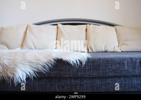 Graues Sofa mit grobem Polstertuch und weißen dekorativen, gewellten Kissen. Über den Sitz wird ein weißer Kunstpelzteppich oder ein Überwurf geschleudert Stockfoto