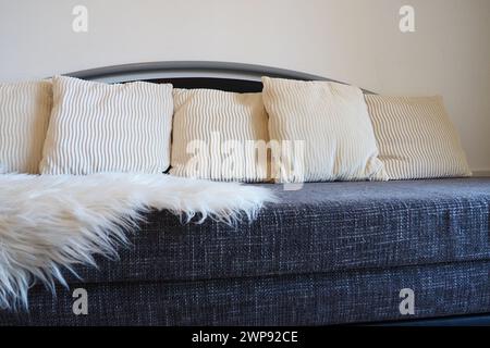 Graues Sofa mit grobem Polstertuch und weißen dekorativen, gewellten Kissen. Über den Sitz wird ein weißer Kunstpelzteppich oder ein Überwurf geschleudert Stockfoto