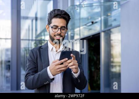 Ein junger indischer Geschäftsmann in einem Anzug ist mit seinem Smartphone verlobt und lächelt, während er eine Nachricht liest oder schreibt. Dieses Bild fängt einen Moment des modernen Berufslebens ein, möglicherweise während einer Pause oder auf dem Weg zur Arbeit. Stockfoto