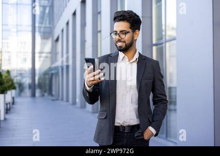 Ein glücklicher und erfolgreicher junger Indianer in formeller Kleidung ist mit seinem Smartphone beschäftigt und lächelt, während er in einer modernen Stadtumgebung steht. Dieses Bild zeigt das Wesen von Konnektivität, urbanem Leben und beruflichem Erfolg. Stockfoto