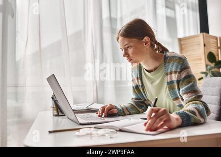 Konzentrierter Teenager, der zu Hause Hausaufgaben macht, Gen z Girl Notizen auf Notebook in der Nähe eines Laptops macht Stockfoto