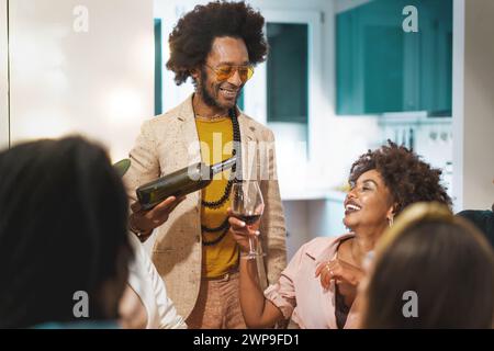Mann im Retro-Afro-Stil serviert Wein für lachende Freunde bei einem intimen Treffen im Haus - Momente des Genusses und der Zweisamkeit. Stockfoto