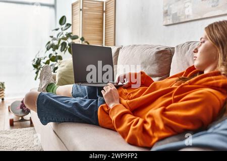 Teenager-Mädchen, das auf einem Laptop mit leerem Bildschirm Filme ansieht und sich auf einem Sofa im Wohnzimmer entspannt Stockfoto