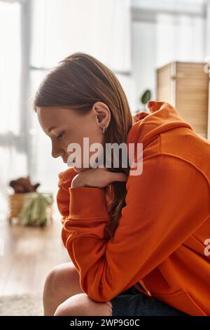 Verärgertes Teenager-Mädchen in orangefarbenem Kapuzenpulli, das auf der Couch in der Wohnung von Moden sitzt, Einsamkeit und Traurigkeit Stockfoto