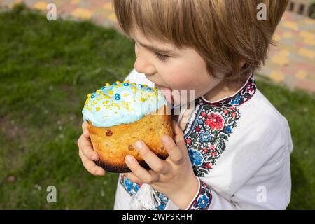 Der Junge schmeckt das süße Sahnehäubchen aus dem Paska. Ein ukrainisches Kind in einem gestickten Kleid hält einen Osterkuchen in den Händen und leckt ihn mit der Zunge. E Stockfoto